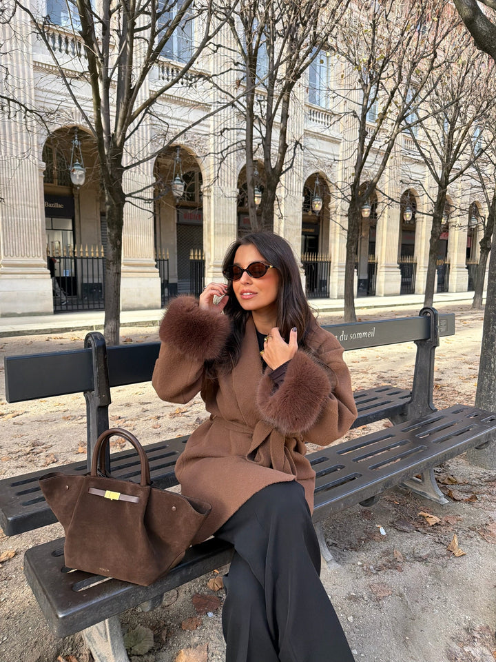 Woman wearing a brown long coat with fur-trimmed cuffs and tie belt