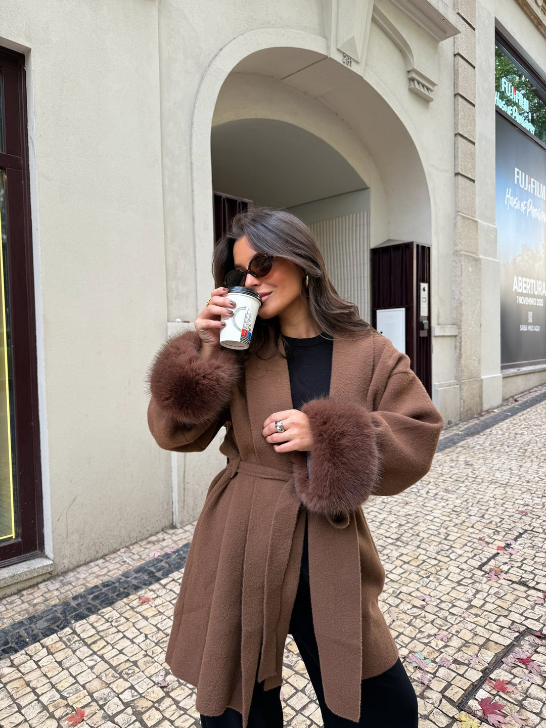 Woman wearing a brown long coat with fur-trimmed cuffs and tie belt