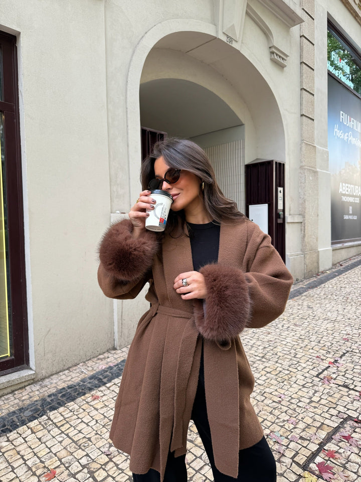 Woman wearing a brown long coat with fur-trimmed cuffs and tie belt